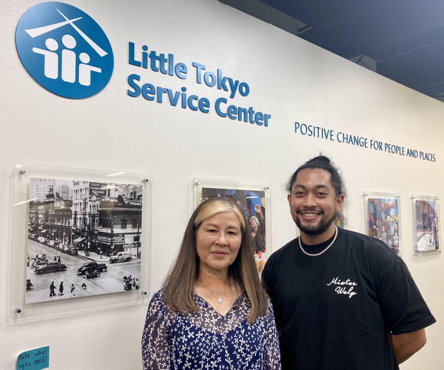 Marian Sunabe and Matthew Yonemura stand side by side inside the Little Tokyo Service Center in Los Angeles. A sign above their head reads "Little Tokyo Service Center: Positive Change for People and Places." They stand in front of a wall of framed photos. The photo to their left is a black and white shot of a street scene from days past. 