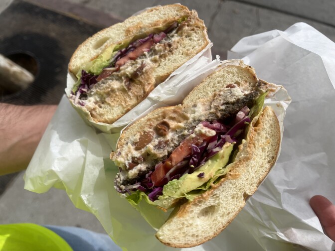 A sandwich, sliced in half to show the insides, is filled with avocado, lettuce, a large slice of red tomato, and pieces of purple cabbage. It sits on top of a thick, crumbly white substance at the bottom of a substantial roll-type bun.