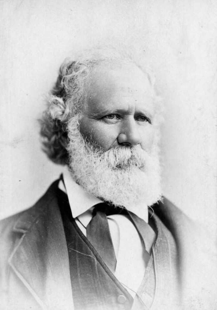 A man with light-toned skin and full white beard wears a suit and vest.