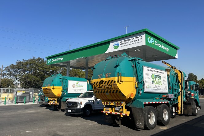A wide shot of green trash trucks refueling at a CNG station. The shade platform over the fueling station is green and reads "clean energy"  and "Los Angeles County Sanitation Districts." It's sunny under a clear blue sky. 