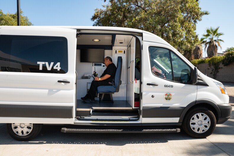 Community health worker Martin Escobedo prepares for an emergency call as part of the Therapeutic Transportation Van program for mental health emergencies across Los Angeles, CA. Escobedo sits in a white van, the side door is open and he appears to be working with a mobile computer device.