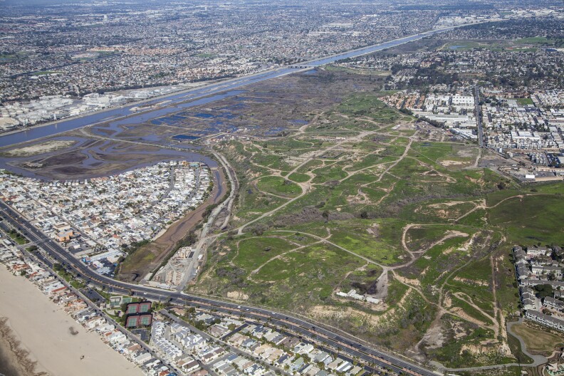 An aerial view of a large, open piece of land crisscrossed with dirt roads. Across the top left corner, you can see a channelized river and beyond that housing developments.
