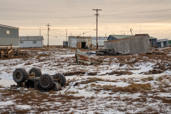 An ATV in the foreground is flipped and partially covered in snow. A home is flipped over and other buildings are damaged in the background.