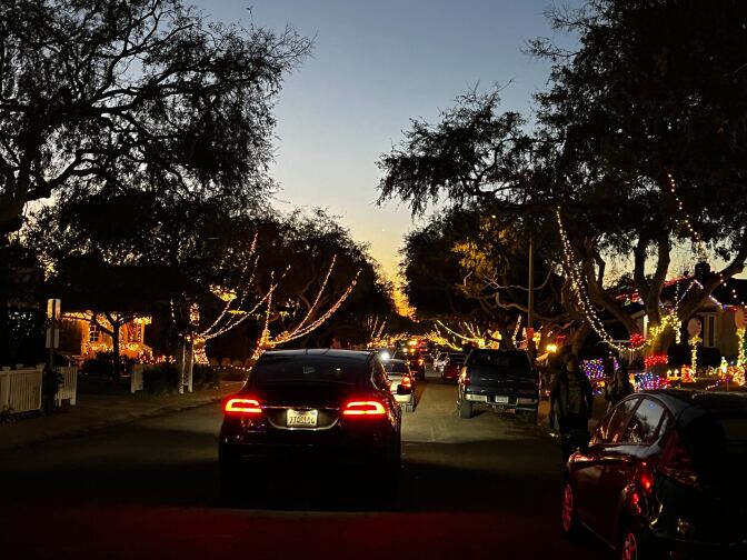Several cars are driving slow down a street, with the houses and trees on each side covered in colorful string lights. The sky is light blue and orange as the sun sets.