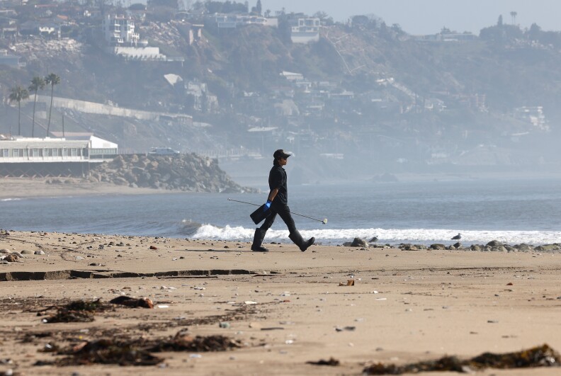 A City of Los Angeles Sanitation worker walks past the tiny bits of pulverized fire debris on the sand at a beach. Homes and greenery can be seen on a hillside in the background.