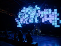 People sit in a dimly illuminated space, on wood benches, looking up at a faux tree. It's made from a real tree's trunk, with digital colored screens in square pixelated shapes creating a faux canopy. The panels at the time of this image are illuminated in light blue and purple.