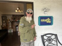 A man with a long, white beard, wearing an army green sweatshirt, sunglasses and a backwards camo-print baseball hat, stands in front of an open doorway with two fingers pointing toward an adjacent sign that reads "micah's way bridging the gaps."