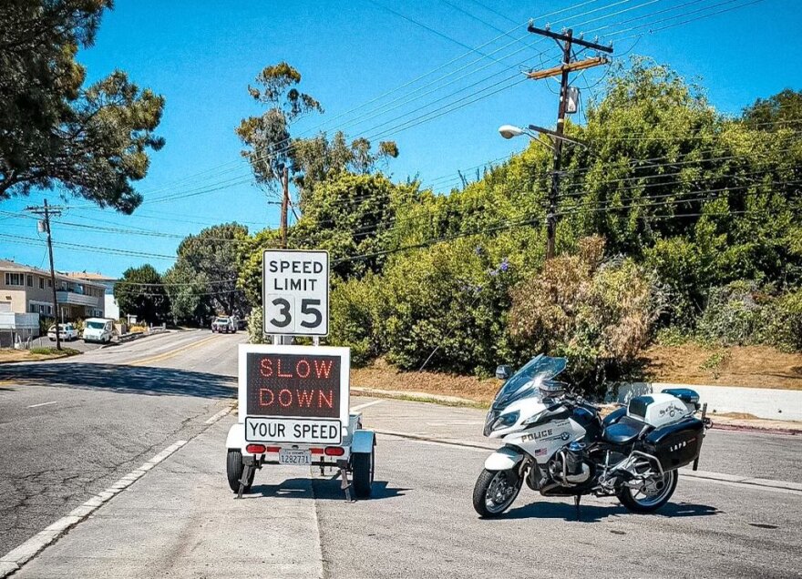 A speed feedback sign stationed on a street next to a police motorcycle shows a speed limit of 35 miles per hour and reads "slow down."