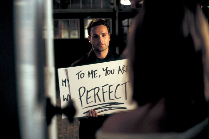 A man holds up a white cardboard poster with the words, "to me, you are perfect." 