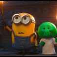 A still of an animated movie, a pill shaped yellow creature wearing overalls holds up a torch, two more creatures like him are in the background. A green smaller creature holding a torch is also present.