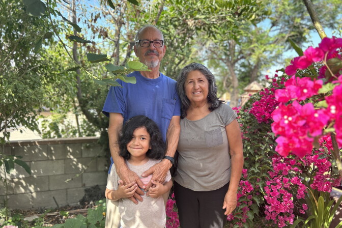 An older male and female couple with brown skin stands with a young boy with long black hair, all smiling for the camera. The man wears a blue t-shirt and the woman a light grey t-shirt. The boy wears an offwhite t-shirt. They are surrounded by trees and bougainvillea. 