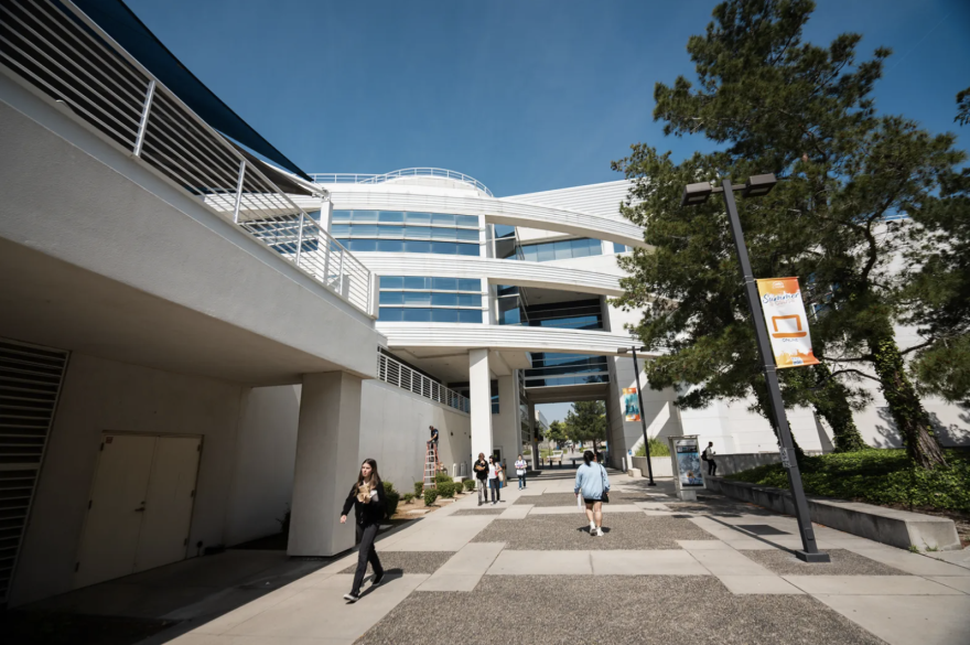 Students walk on a college campus during the day, large white buildings looming over them.