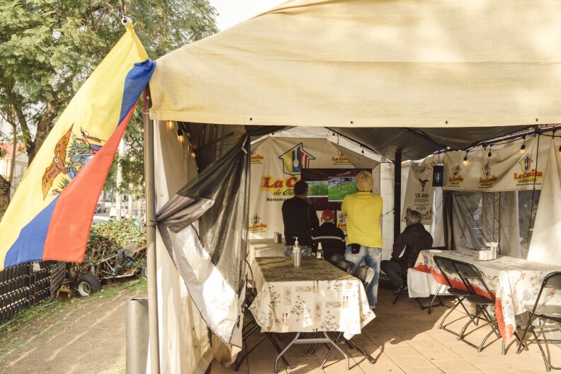 Tables in a pop-up tent are covered in tablecloths. People in the back of the tent are watching TV