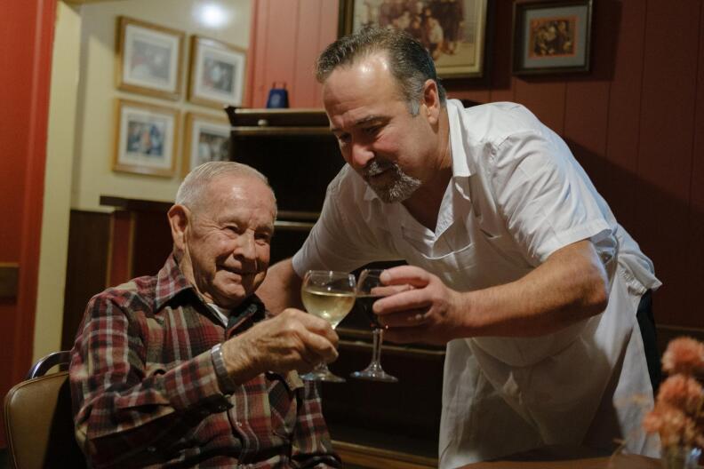 a man in a plaid shirt sits inside an empty dining room clinks his wine glass with another man wearing a chef's white uniform