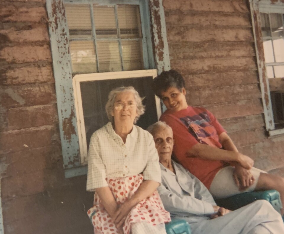 Three people sit together on a front porch of a home with peeling paint.