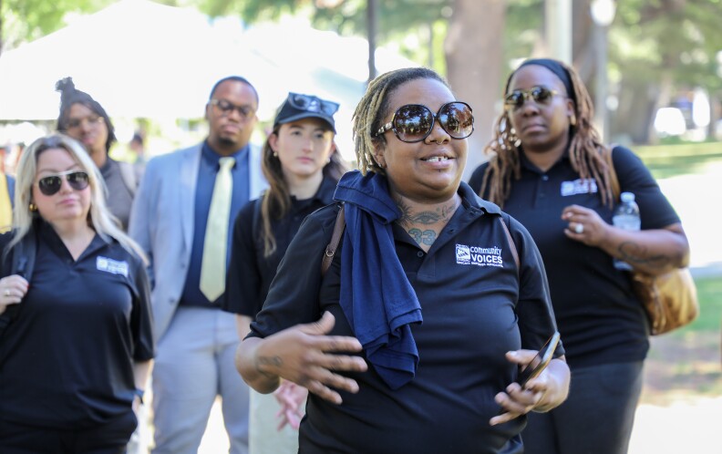 A Black woman with short bleached twists wearing a navy blue Community Voices collared shirt and large sunglasses, stands before a group of people gesturing her hands.