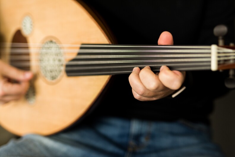 Two fair-skinned hangs holds the oud. In the foreground a set of fingers is wrapped around the neck of the fretless stringed instrument.