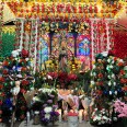 At the center of the altar is a statue of the Lady of Guadalupe -- a brown-skinned woman wearing a green veil with her hands clasped in prayer and an angel at her feet. Behind the statue is a tapestry with a glass-stained window design. The statue is surrounded by flowers of all kinds of colors. 