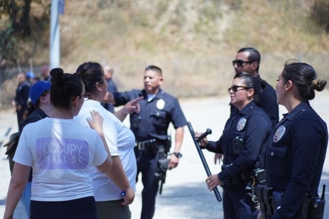 A woman in a white T-shirt speaks to uniformed LAPD officers, one of whom holds a night stick.