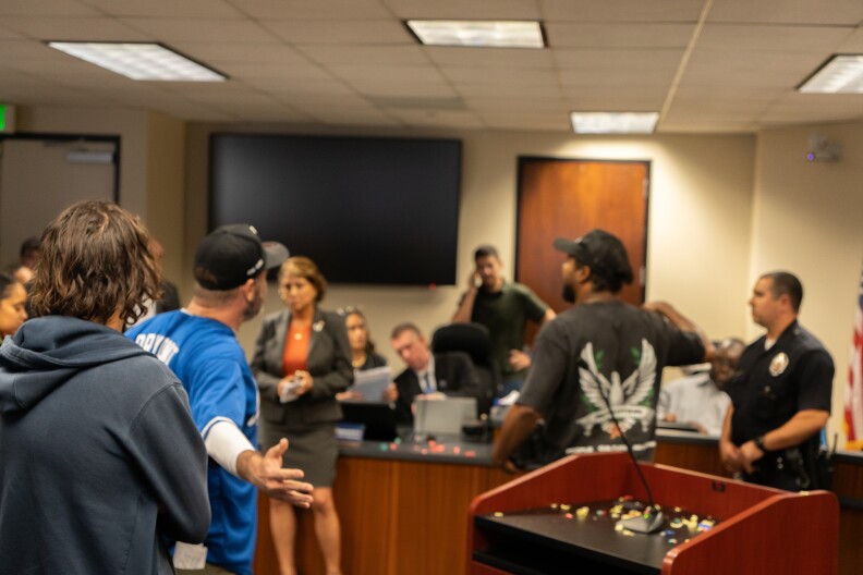 People in a meeting room appear agitated. One man has his arms outstretched and another is gesturing angrily with his right arm.