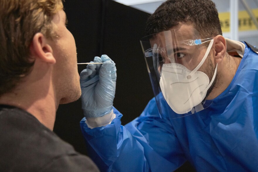 A medical worker wearing blue scrubs, a mask and face shield extends a hand with a swab into a patient's month.
