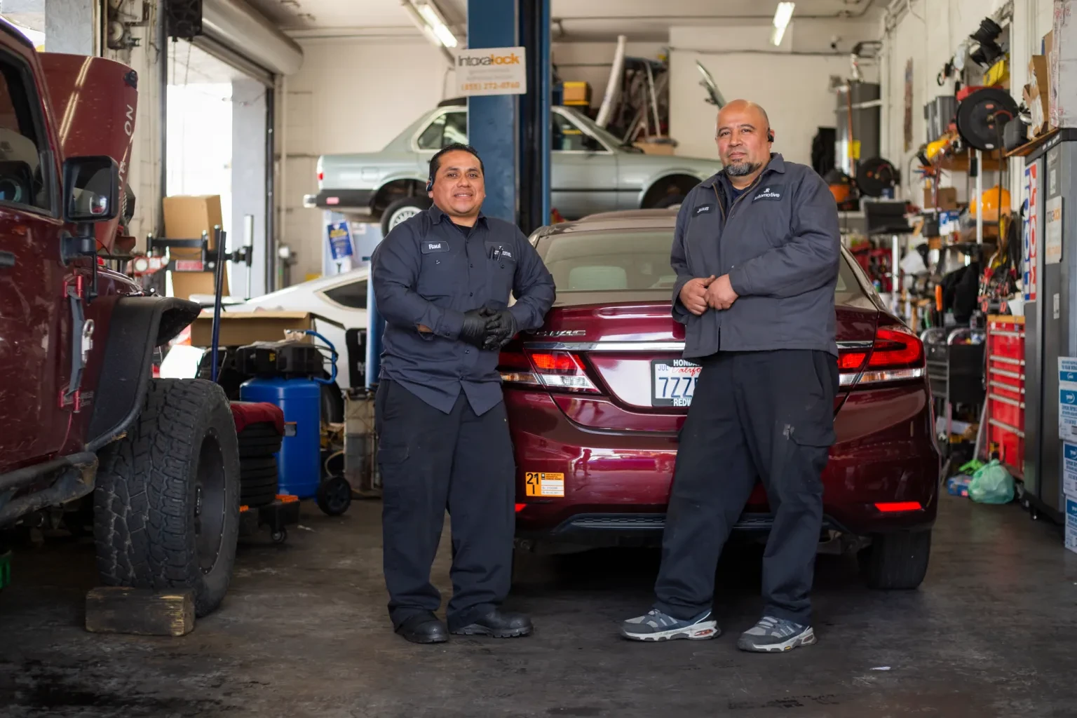 Two men pose in front of a red car inside an auto mechanic garage.