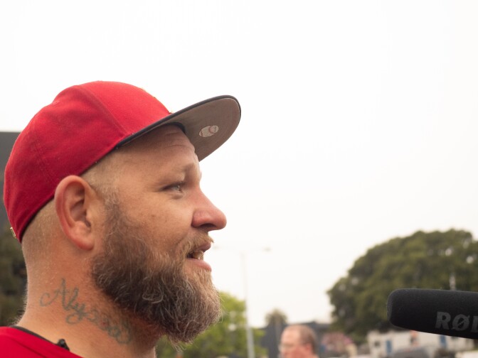 A side profile of fair-haired, bearded man in a red cap and t-shirt with a tattoo that reads "Alyssa" in cursive on his neck, speaking into a microphone