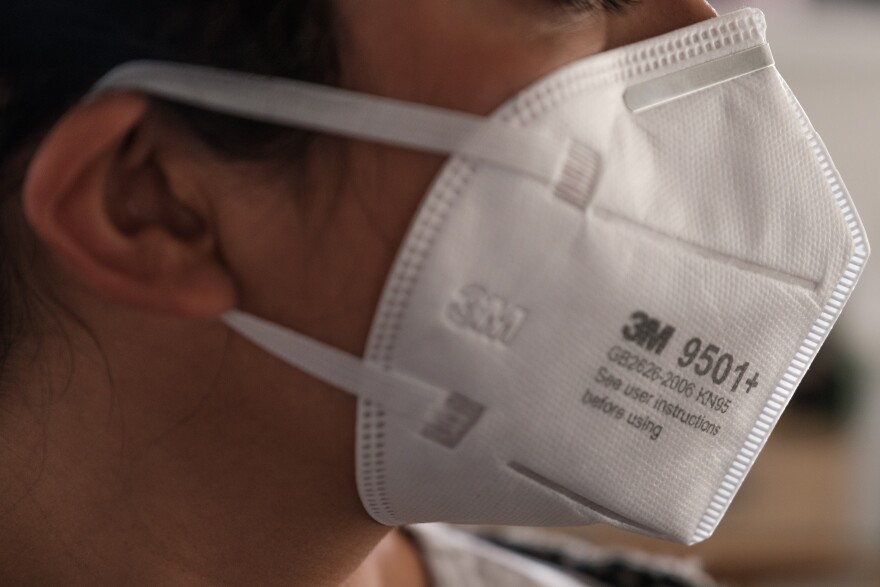 A close-up profile of someone wearing a white, high-grade medical mask. A white strap is wrapped around the person's right ear.