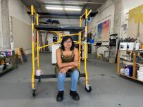 A brown skinned woman sits on a yellow dolly in the middle of a warehouse. 