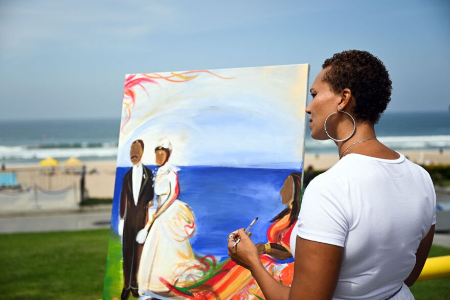 A woman with dark-tone skin holds a paint brush near a canvas on an easel at a grassy beach near the ocean. The paint depicts a Black couple in formal dress with the water behind them.