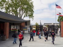 Young kids with backpacks walk through a gate of what looks like a school. It is outdoors and there is a flag pole with an American/California stage flag