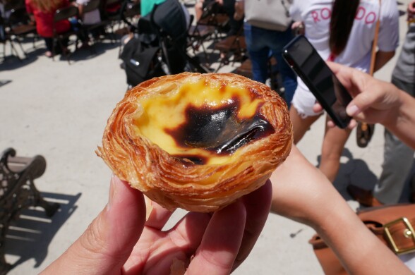 A hand is holding a round custard tart with light brown flakey edges, the custard is in the center and has an oblong black spot on top from being burned.