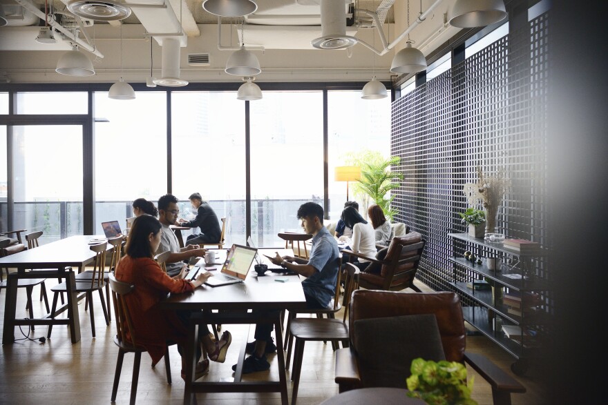 People sit at tables and on lounge chairs in a well-lit room with exposed ducts.