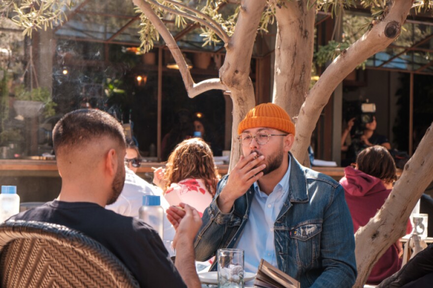 A man wearing an orange beanie sitting across a table from another man smokes a marijuana cigarette at The Original Cannabis Cafe in West Hollywood. Other patrons are seen in the background at the restaurant that allows patrons to partake of cannabis on the premises.