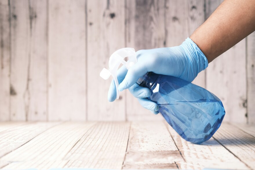 A stock image of someone with a blue glove spraying a blue bottle onto a wooden floor. 