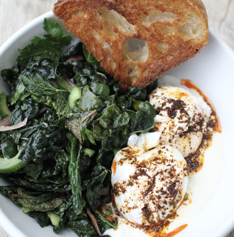 A white bowl contains sauteed greens, two softly poached eggs sprinkled with spices, and a piece of toasted rustic bread