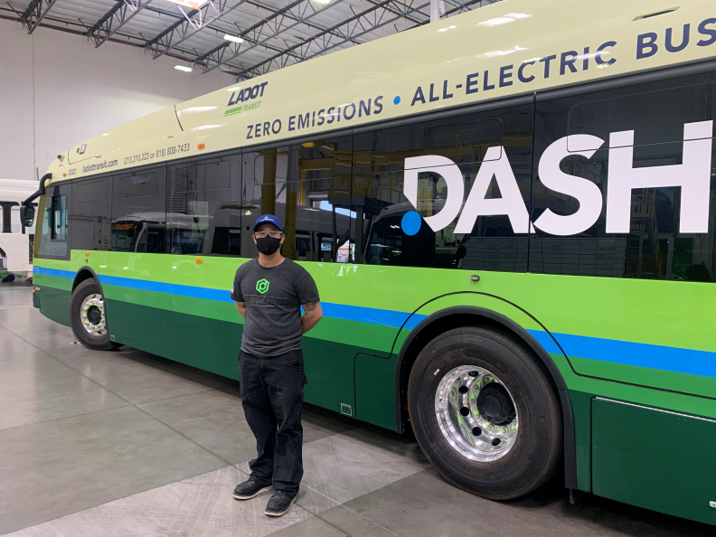 Albert Baena standing with face mask on, hands behind back, in front of an electric bus at the Proterra manufacturing facility in the City of Industry.
