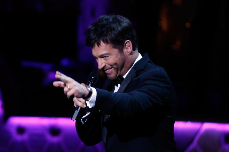 Singer Harry Connick Jr. performs on stage in a suit, smiling and gesturing to the audience. Behind him, the dark stage is bathed is accented with purple light.