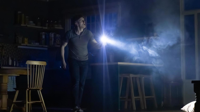 A man holds a flashlight in a dimly lit environment, surrounded by a set that appears to be a kitchen.