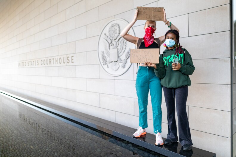 In front of a sign for the United States Courthouse, two people with masks on hold signs up. The clearest one reads keep your bans off my body.