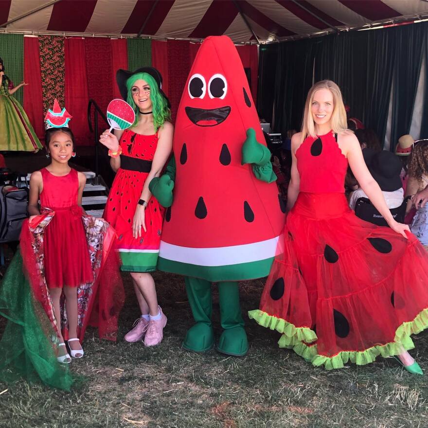 Four people dressed in watermelon-themed costumes stand under a tent decorated with red and green fabric. The first person, a young girl, wears a red dress with green and red accent. The second is a woman wearing a red dress with black seeds and green hair, holding a watermelon-shaped fan. The third is in a large watermelon mascot costume with green gloves and shoes. The fourth is a woman in a red dress with black seeds and green trim, holding out the skirt to display the design.