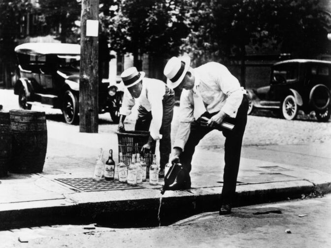 circa 1920: Two men pouring alcohol down a drain during prohibition in America.