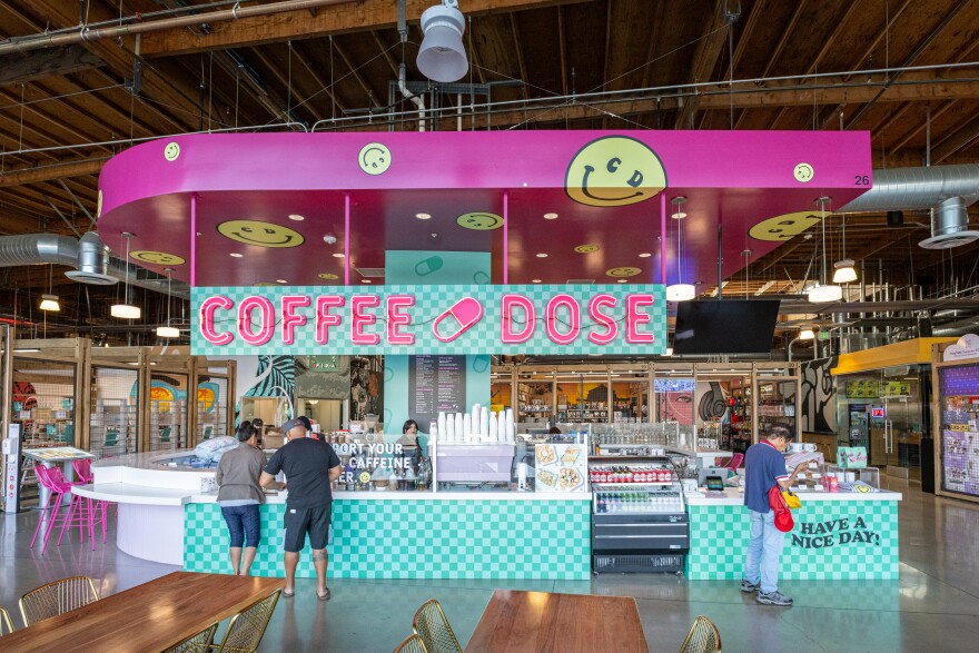 A large pink and teal checked food stand says coffee dose in the middle, amid yellow smiley faces. it's a coffee bar and three people stand in front of it. The stand is in a cavernous open indoor location