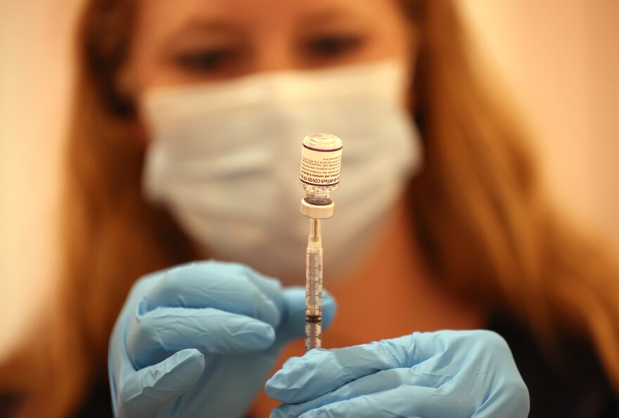 A woman wearing a surgical mask and blue gloves holds a syringe that is inserted vertically into an upside-down vial. The label indicates that it is the Pfizer COVID-19 vaccine.