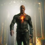 'The Rock' Johnson stands in the center of the image, mid-stride. He wears a skin-tight suit that is dark gray and covers his chest, arms, and down through his legs. The suit has a cracked texture, like lava rock or charred wood. A large, bulky leather belt sits around his waist, the same color of the suit, but a smoother texture. In the middle of Johnson's chest is a large orange lightening bolt that glows as if made of lava. Johnson stares straight ahead, eyes hooded. Behind him is an elaborate hallway with intricate wall designs of gold, emerald, and blue.