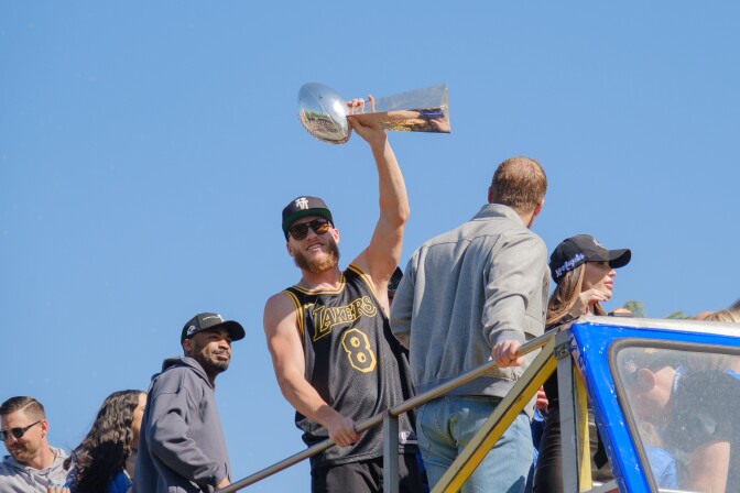 Cooper Kupp holds the Super Bowl trophy in the air and smiles. He wears a black Kobe Bryant jersey. There are a handful of people standing around him also smiling.