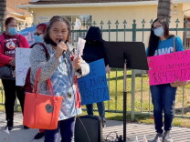 A woman of Asian descent with a gray bob speaks into a microphone on a sidewalk outside the fenced yard of a stucco house. Several other women stand in the background with signs. One pink sign reads "Pay up!"