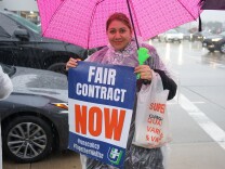 A person with medium-light skin tone holds a pink umbrella and a sign that reads "Fair Contract NOW." Behind her cars are driving in traffic.
