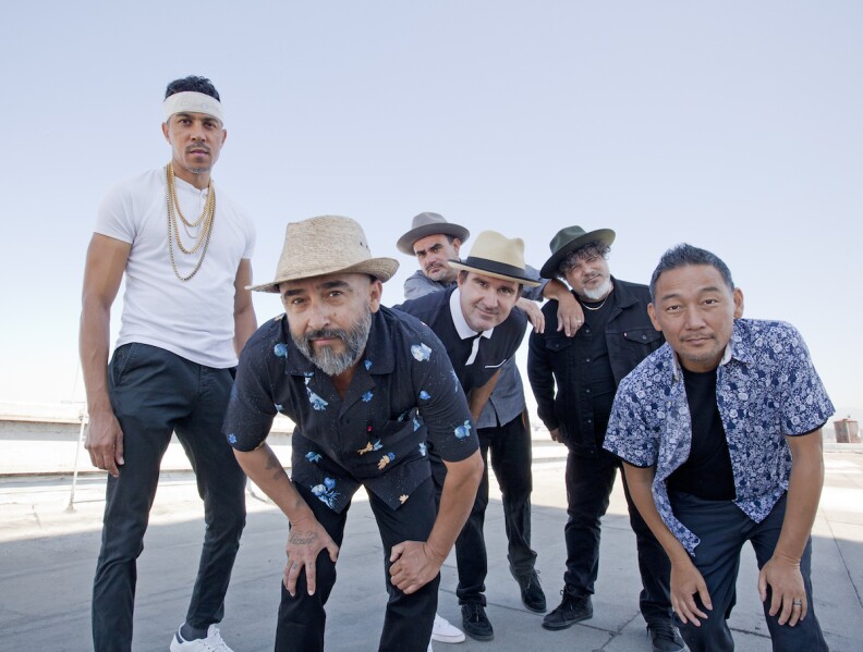 The members of the band Ozomatli bend toward the camera. Four wear hats and one wears a headband and chains. 