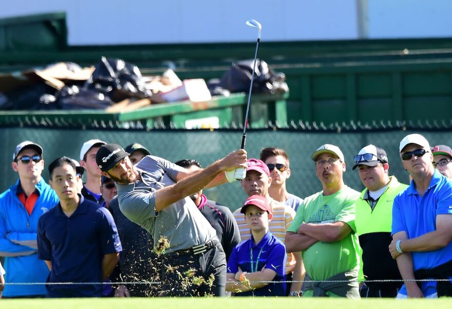 Dustin Johnson in a grey golf shirt and black cap hits a golf shot as spectators watch.